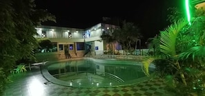 A nighttime view of a swimming pool area with a checkered floor, surrounded by palm trees and a building with illuminated windows.
