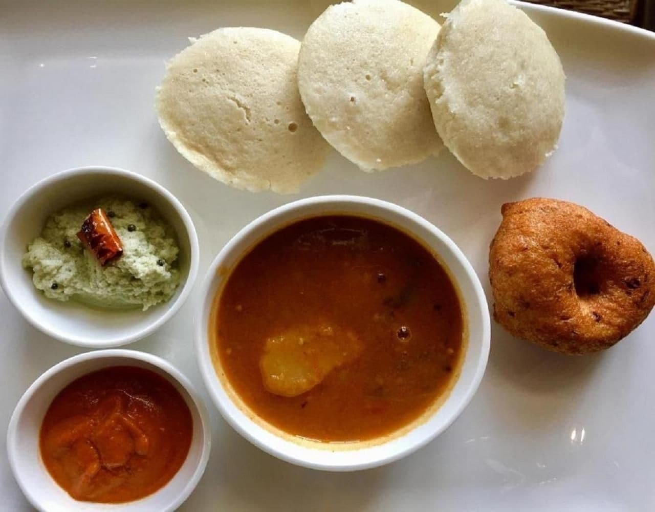 A plate with three idlis, two chutneys, a bowl of sambar, and a fried snack.