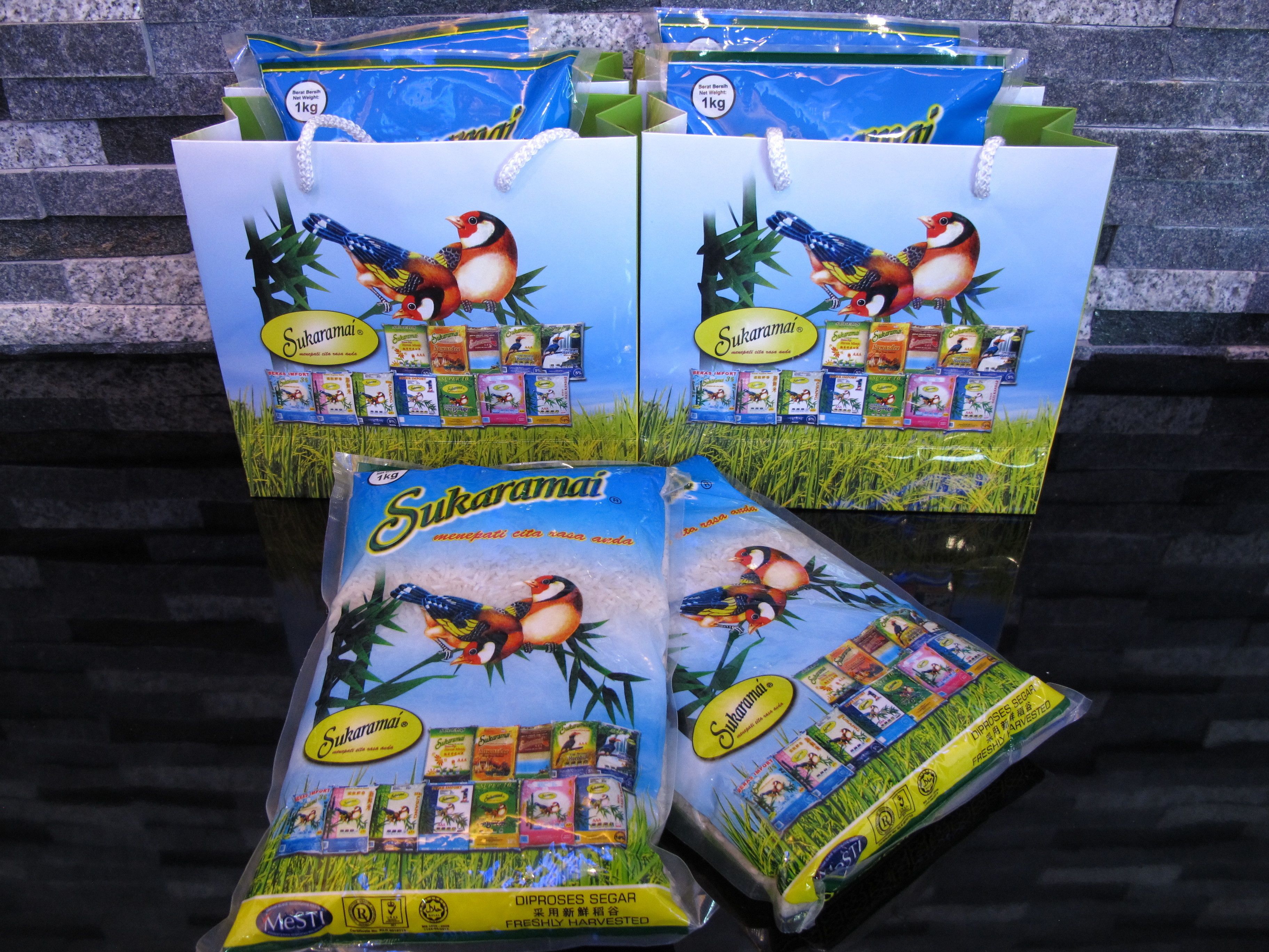 Plusieurs emballages de produits de la marque Sukaramai, notamment du riz et divers produits alimentaires, sont présentés sur une surface réfléchissante, avec un mur de pierres en arrière-plan.