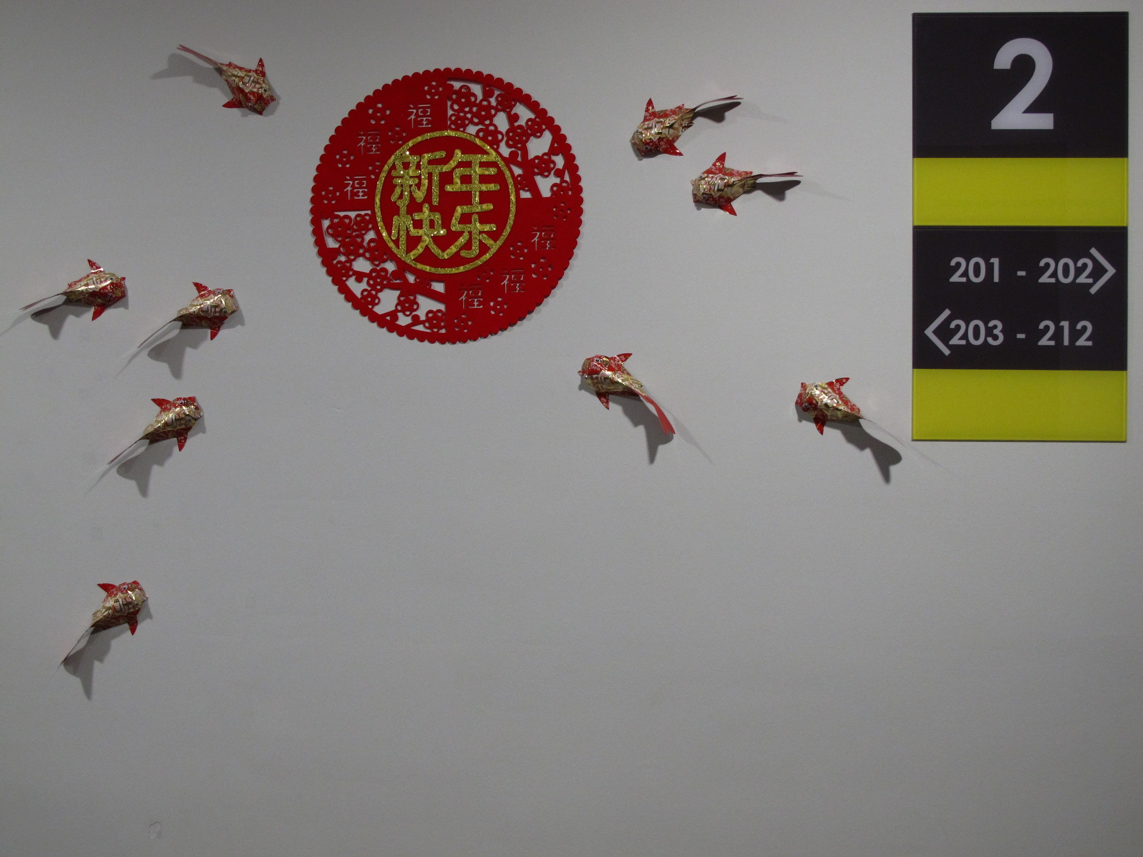A wall with a red circular emblem surrounded by origami fish, and a directional sign with room numbers.