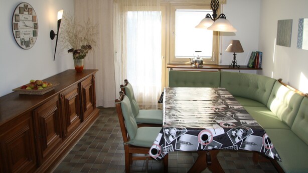 A dining area with a green upholstered sofa, a wooden dining table with a patterned tablecloth, a wooden cabinet with a fruit bowl, and a wall clock. - Gästezimmer Lußhardthof (Sankt Leon-Rot)
