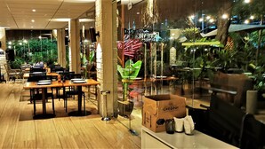Ein Restaurant mit Holztischen und -stühlen, einer Glastrennwand mit Pflanzen und einem sichtbaren Versandkarton auf der Theke.