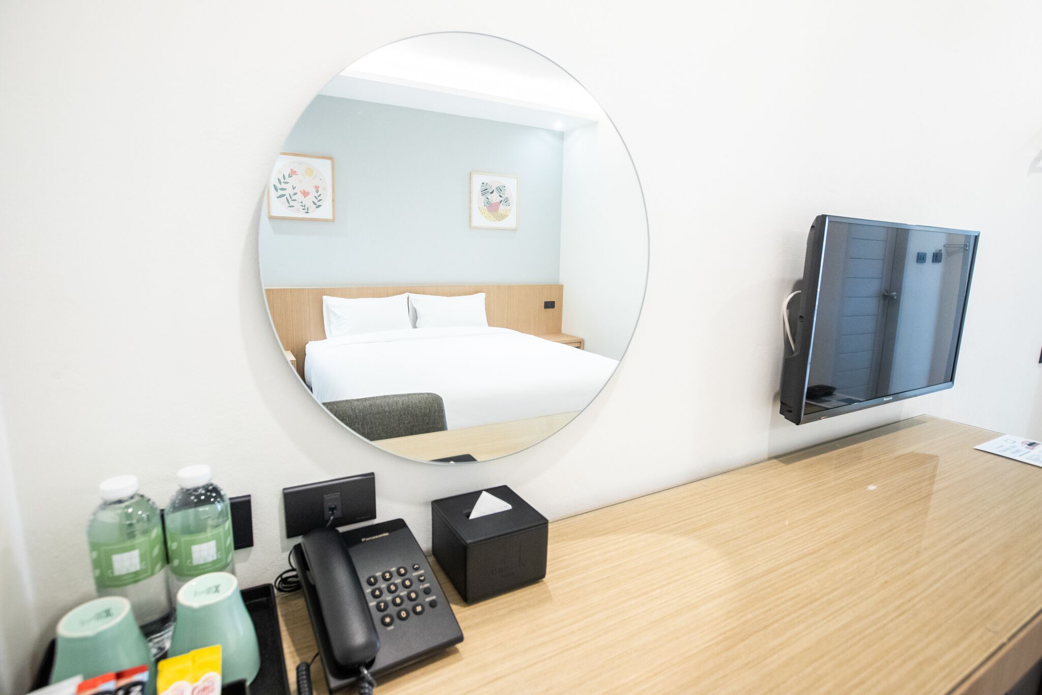 Ein Hotelzimmer mit Bett, Spiegel, Fernseher, Telefon und Schreibtisch mit einer Wasserflasche und einer Schachtel.