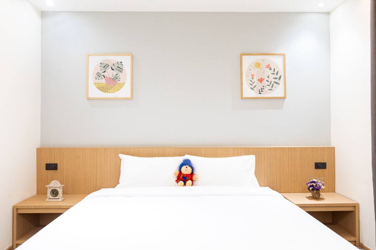 A neatly made bed with a plush toy, two framed artworks on the wall, and a small clock on a bedside table.