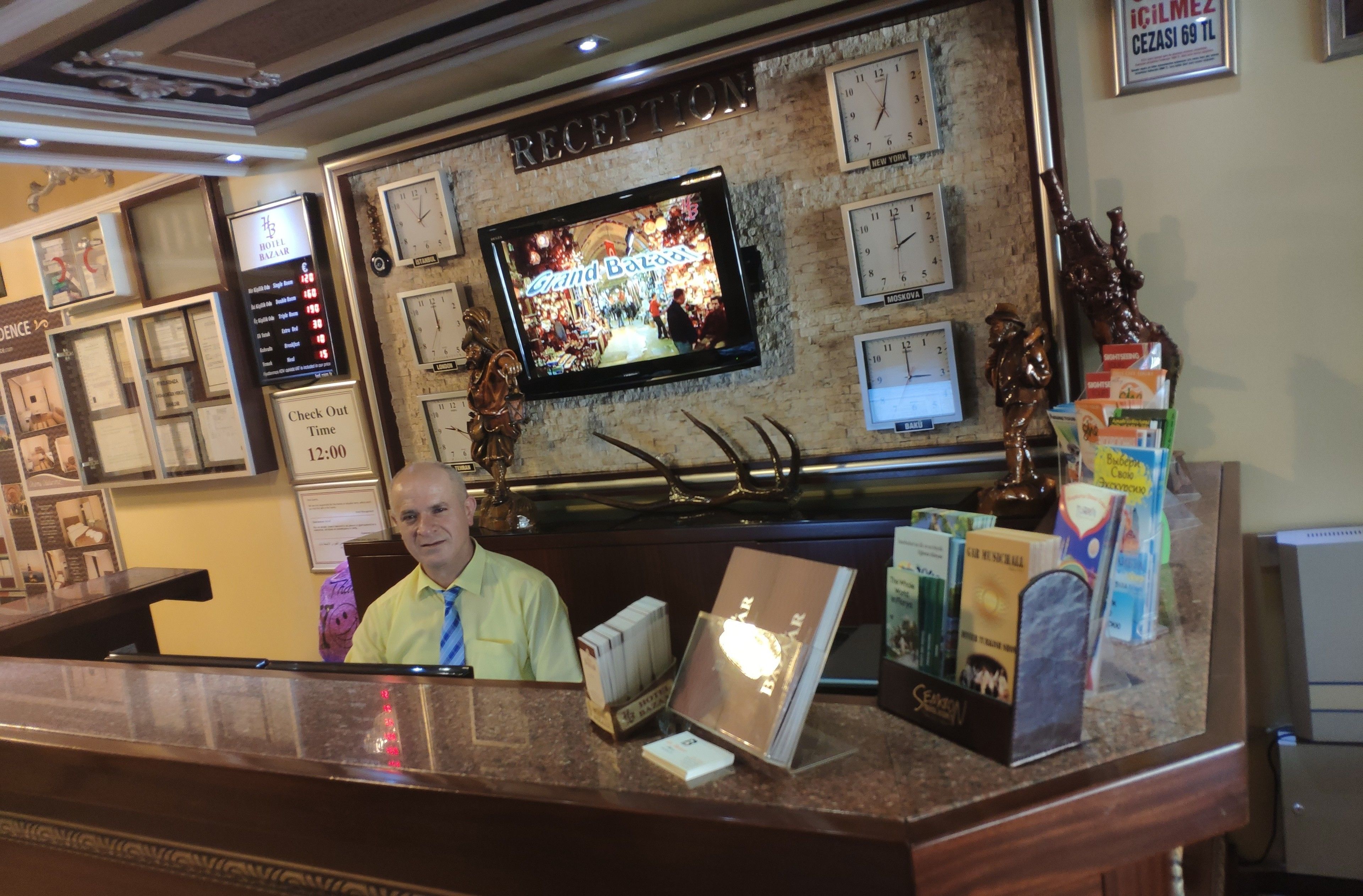 A hotel reception desk with a television displaying 'Grand Budapest Hotel', clocks on the wall, and a 'Check Out Time' sign.