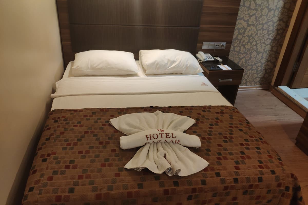 A hotel room with a neatly made bed, a pillow, a bedside table with a phone, and a wall with floral wallpaper.
