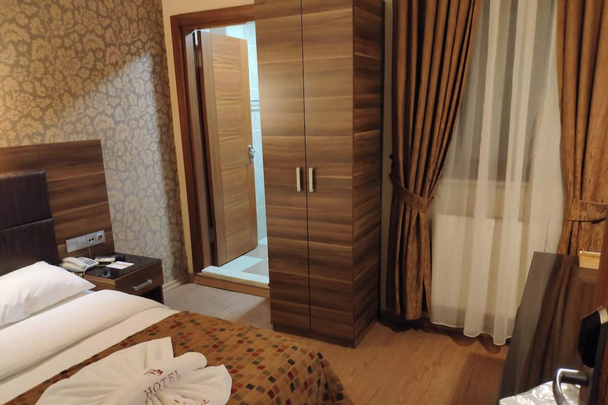 A hotel room with a bed, a wooden wardrobe, a nightstand with a phone, and a bathroom door.