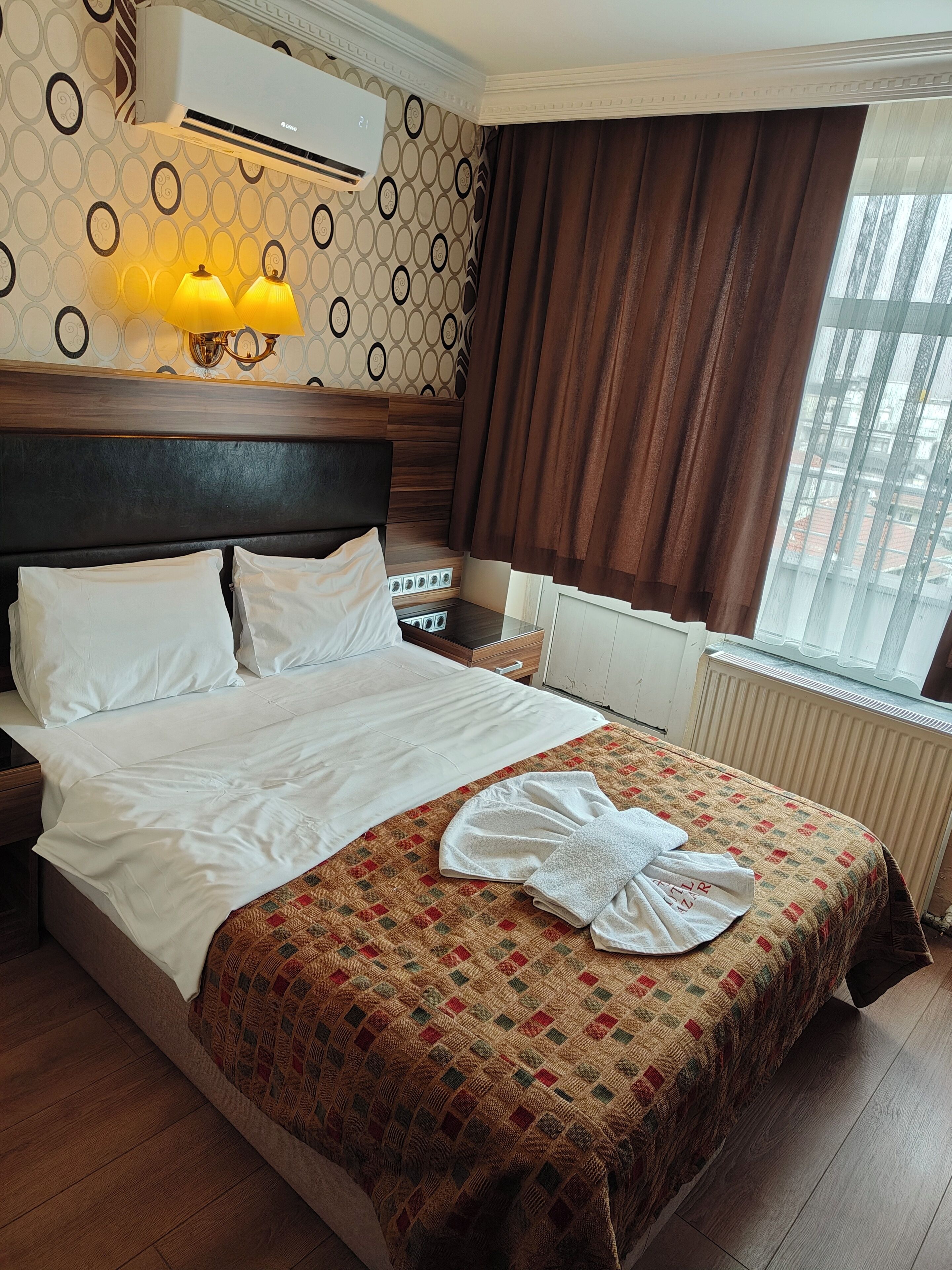 A hotel room with a bed, a bedside table, an air conditioner, a window with curtains, and a patterned wall.