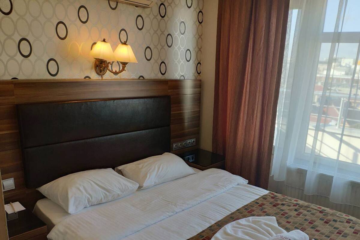 A hotel room with a bed, a nightstand, a wall-mounted lamp, and a window with curtains.