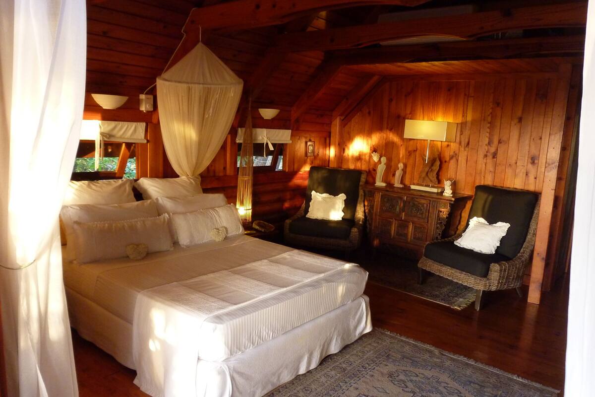 A cozy wooden cabin bedroom with a large bed, two chairs, a nightstand, and a hanging canopy.