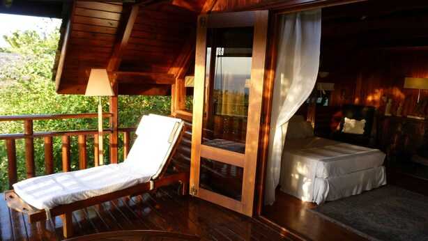 Eine Holzterrasse mit einer Liege, einem Bett mit weißer Bettdecke und einer Glastür, die zu einem Schlafzimmer führt.