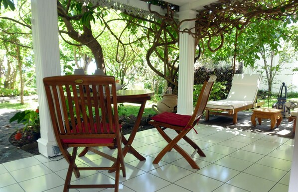 Un patio techado con mobiliario de madera, que incluye una silla y una mesa, rodeado de exuberante vegetación y árboles.
