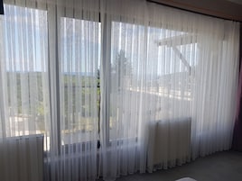 Una habitación con cortinas blancas translúcidas, piso de baldosas y vistas a un jardín y árboles.