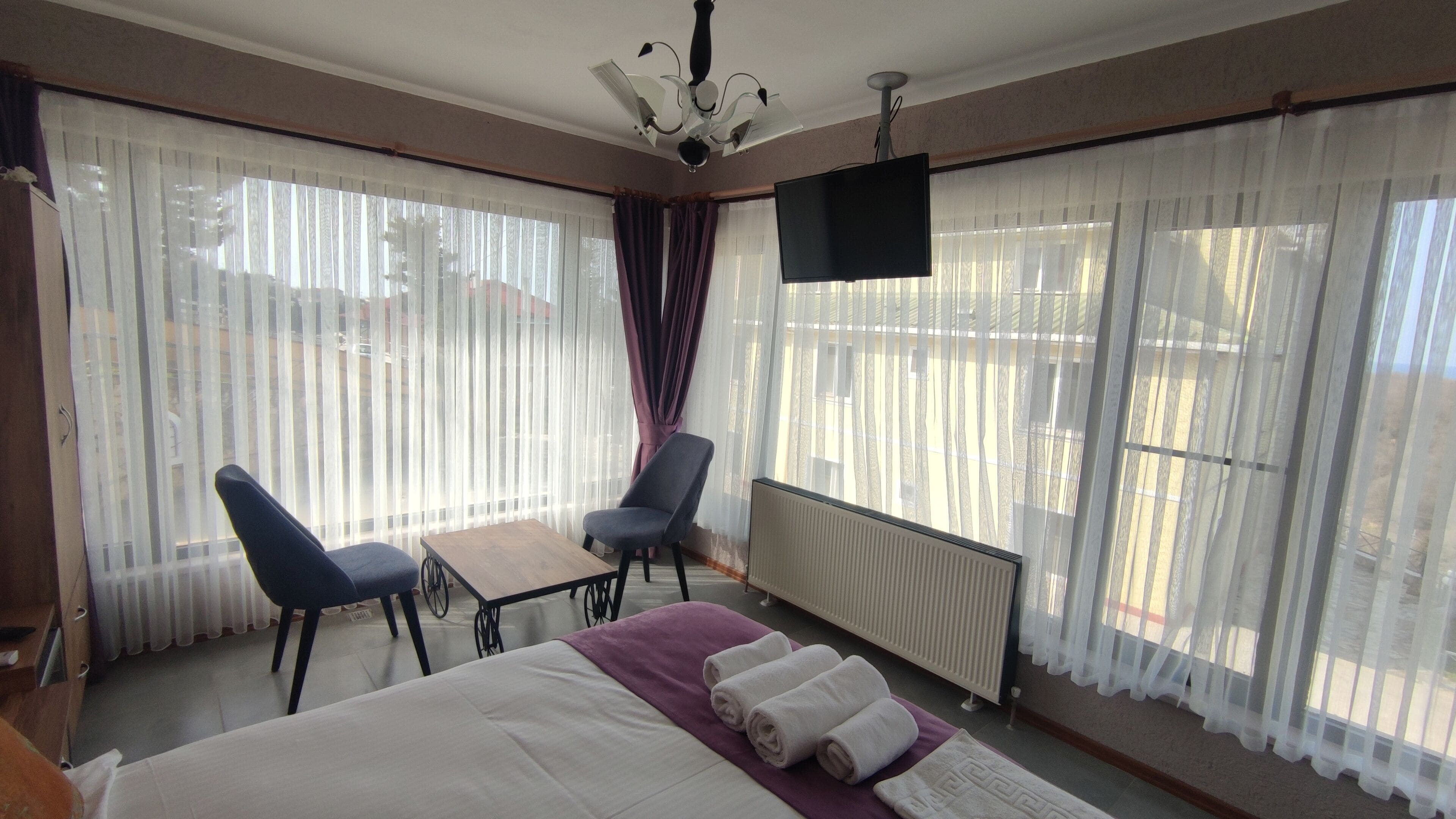 Ein Hotelzimmer mit Bett, Schreibtisch, zwei Stühlen, Fernseher und großen Fenstern mit Vorhängen.
