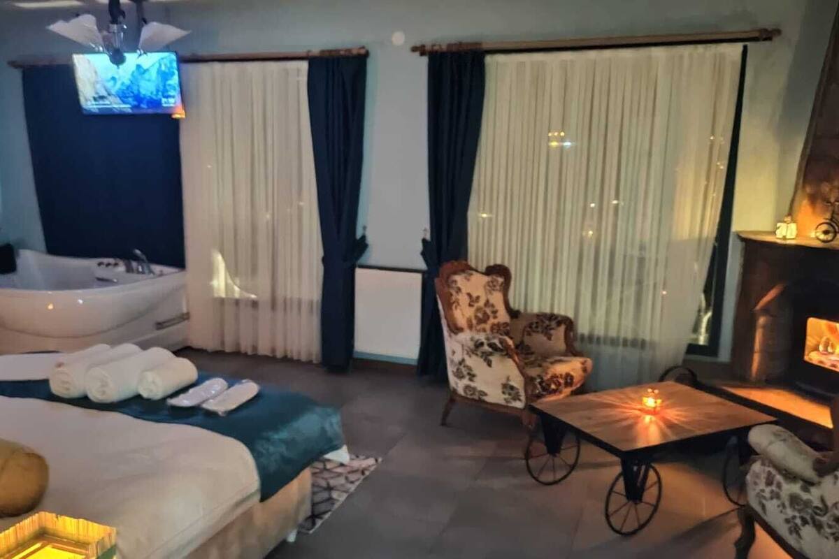 A hotel room with a large bed, a sitting area with a table and chairs, a fireplace, a TV, and a bathtub.