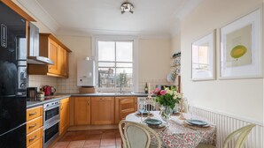 A kitchen with wooden cabinets, a black refrigerator, a tiled floor, a dining table with floral tablecloth, and chairs. - Panoramic Family Home in Maida Vale (London)
