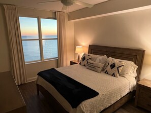 A hotel room with a large bed, a nightstand with a lamp, and a view of the sea through the window.