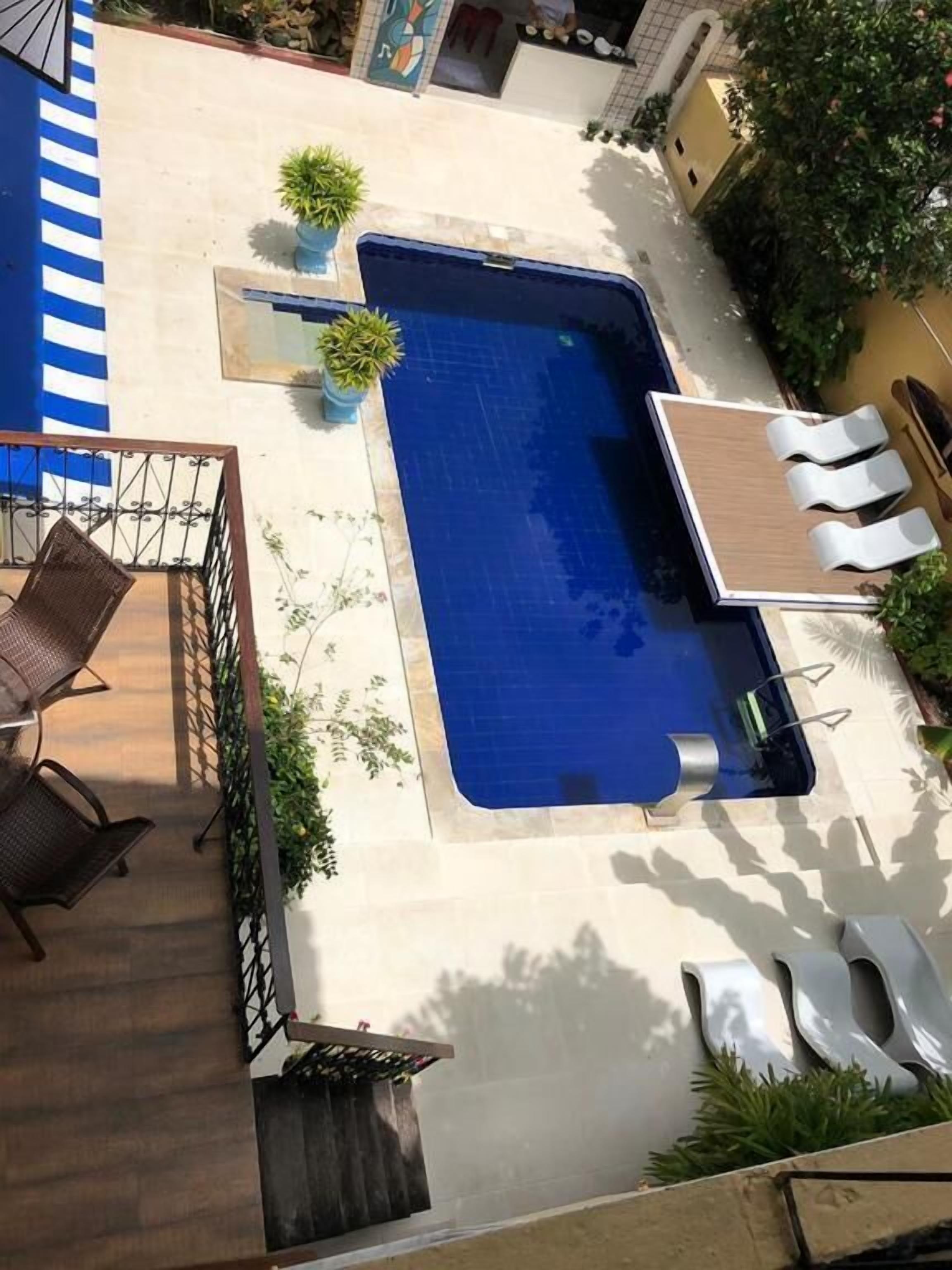 A rectangular swimming pool with a wooden deck, surrounded by potted plants and white lounge chairs.