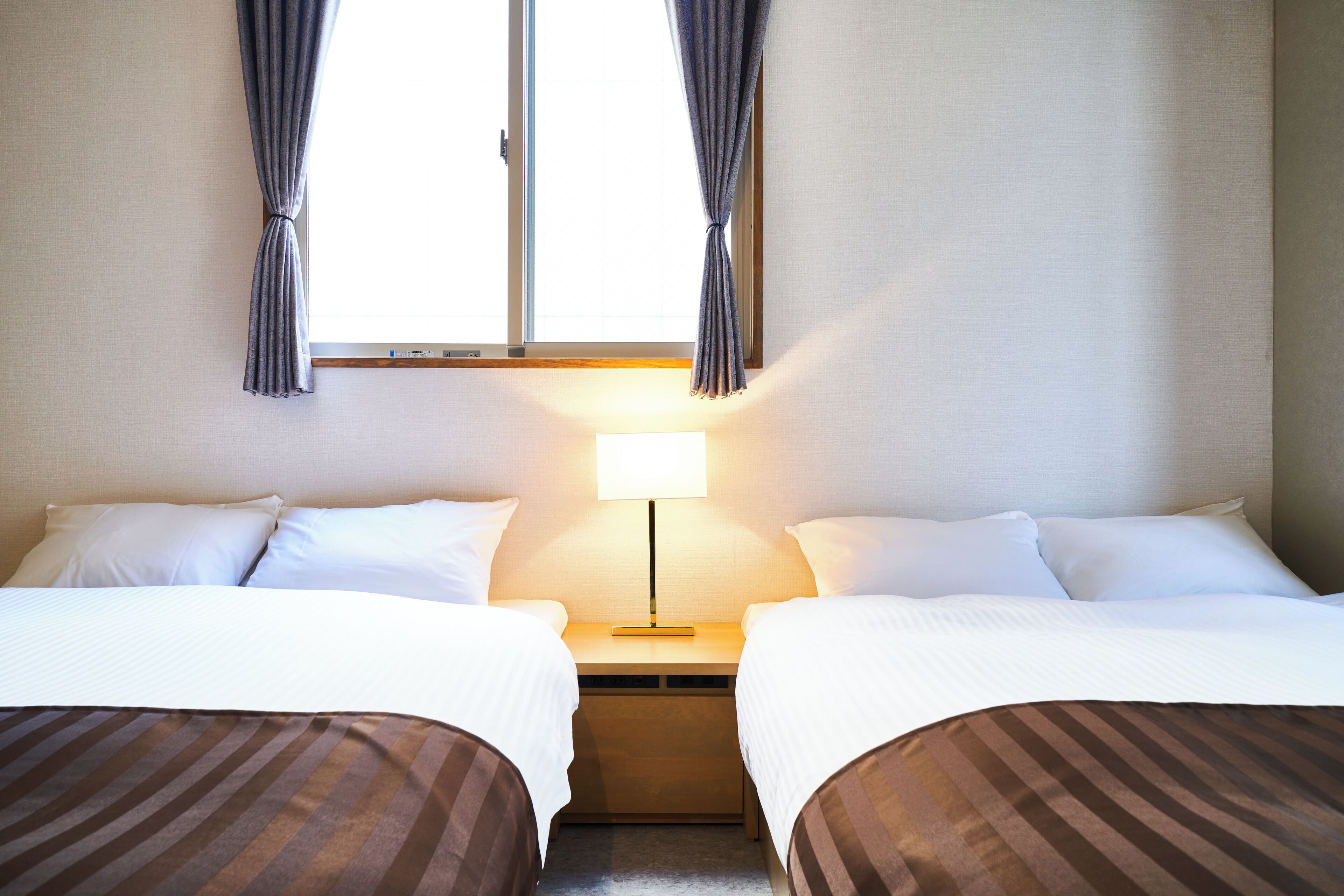 A hotel room with two beds, a nightstand, a lamp, and a window with curtains.