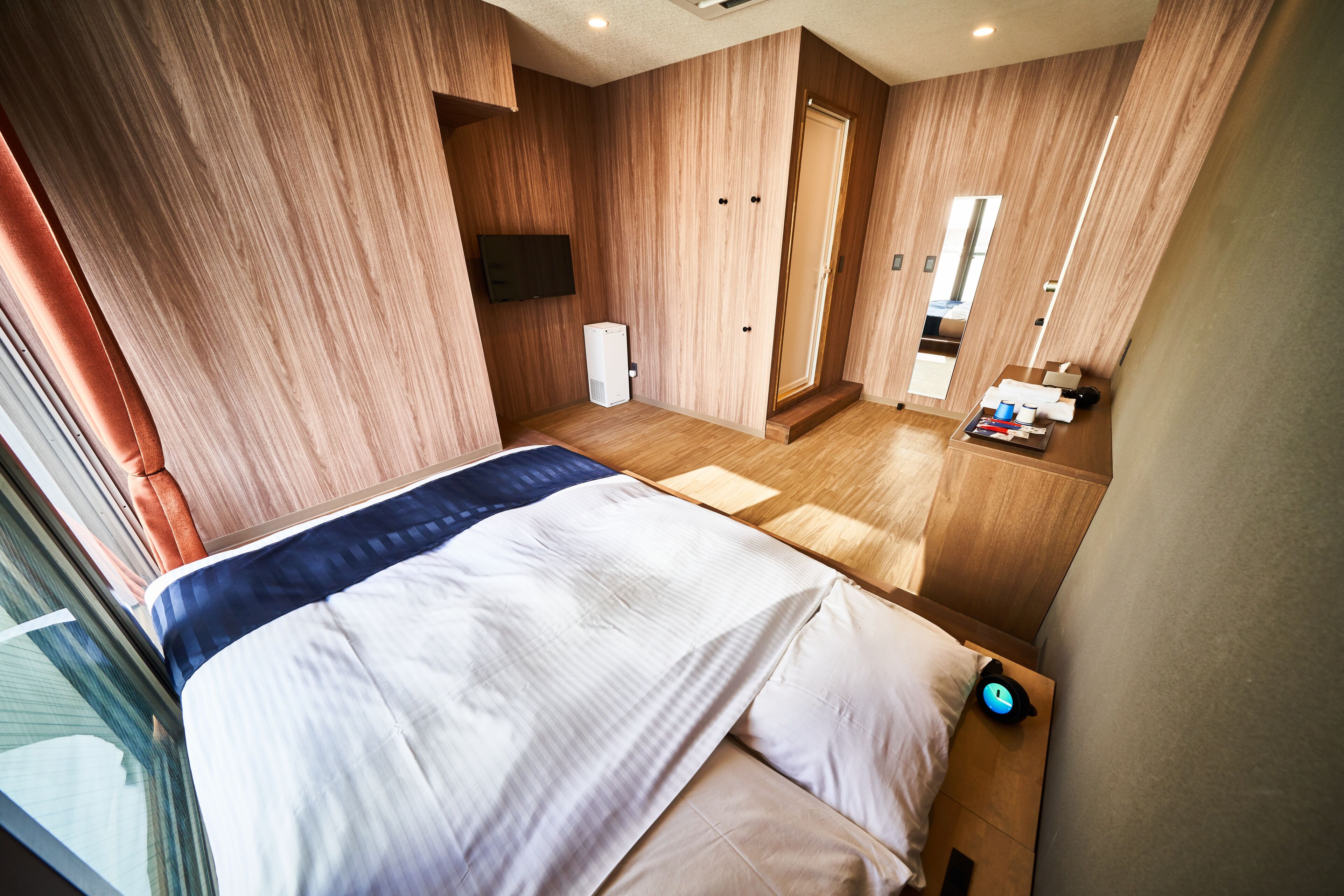 Una habitación de hotel moderna con una cama grande, paredes de madera, un televisor de pantalla plana, una mesita de noche pequeña y un baño con espejo y lavamanos.