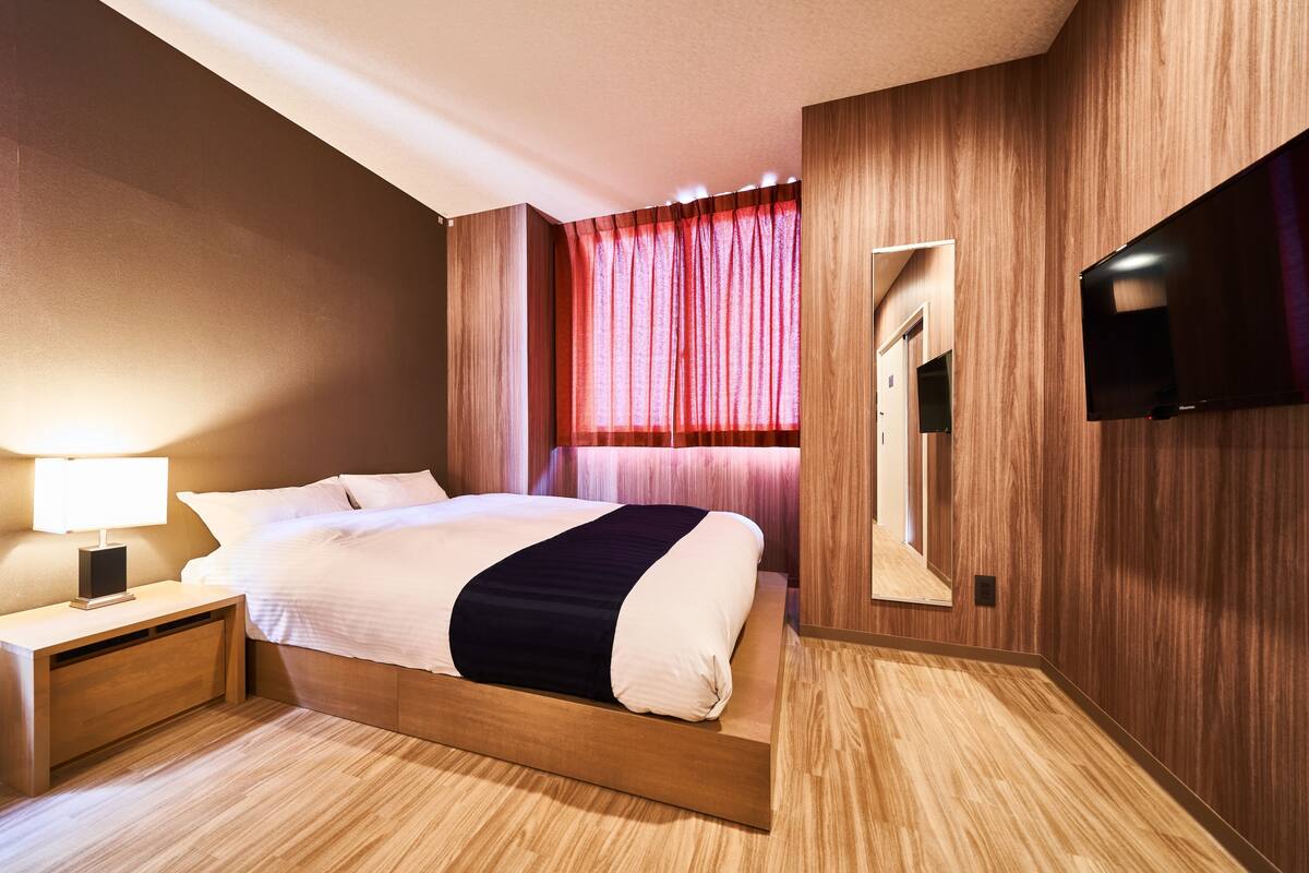 A modern hotel room with a wooden bed frame, a flat-screen TV mounted on the wall, and a nightstand with a lamp.