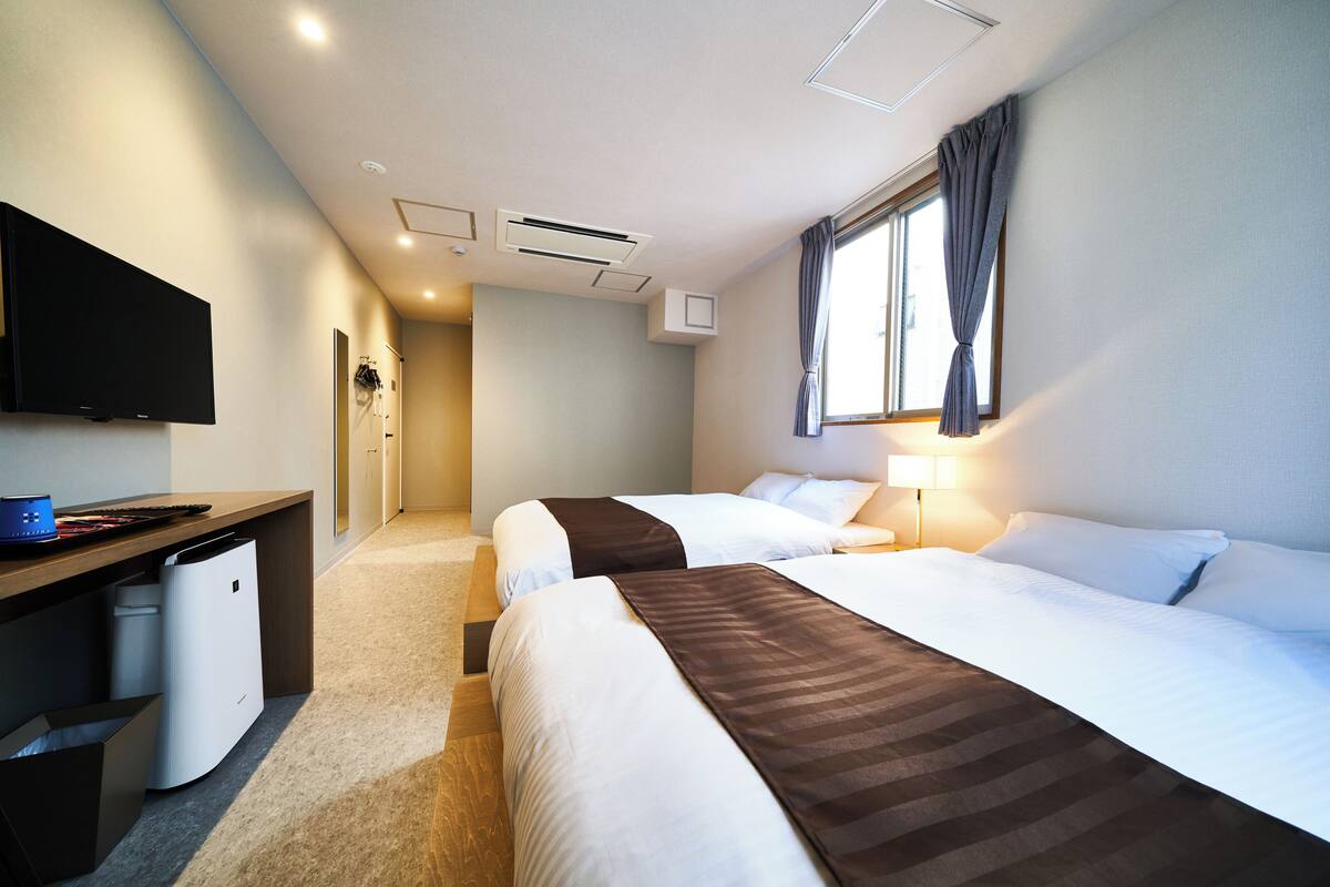 A hotel room with two beds, a desk, a television, and a window with curtains.
