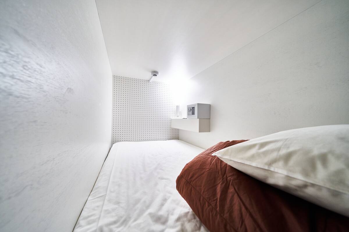 A small, modern bedroom with a single bed, a white wall-mounted shelf, and a ceiling light.
