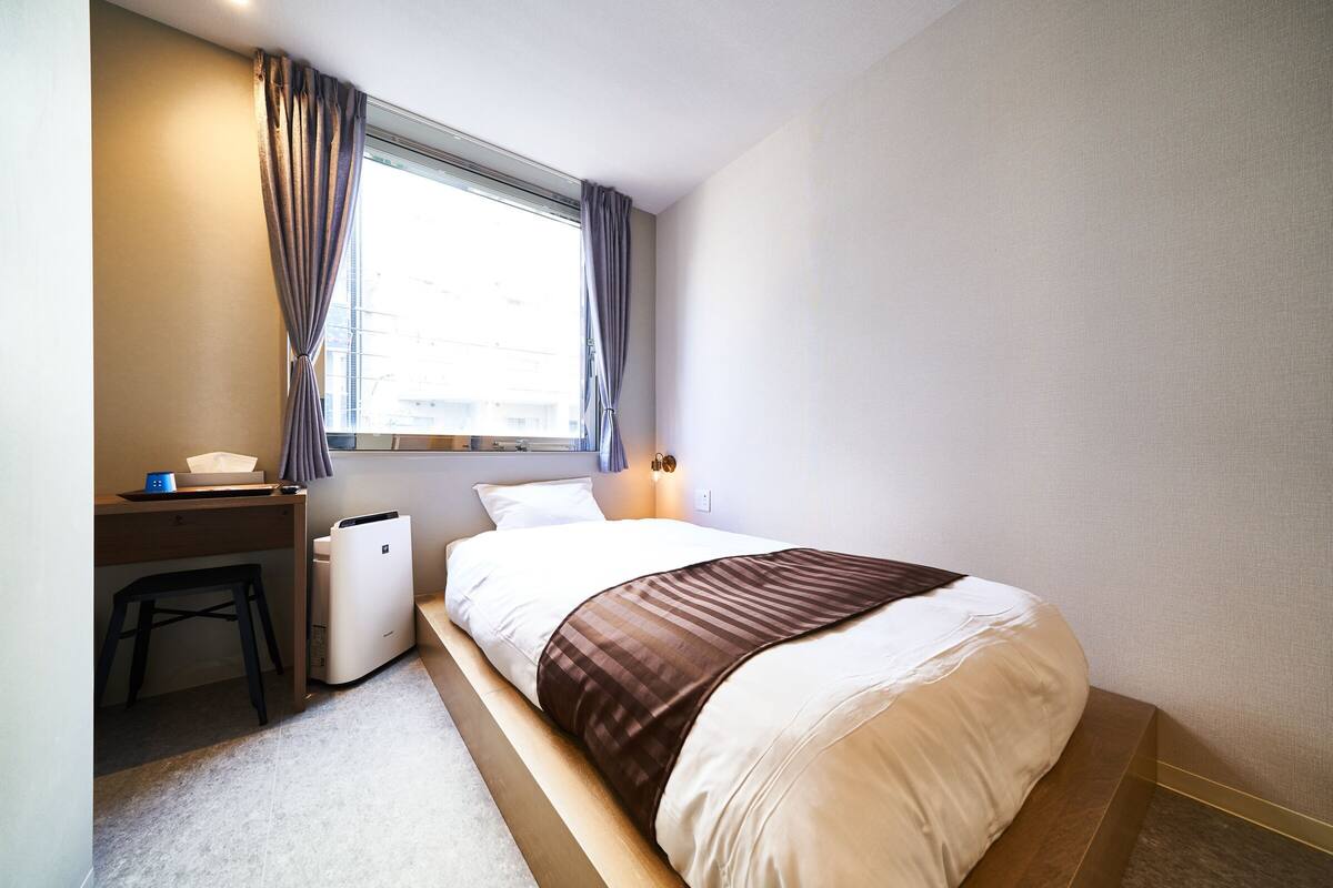 A hotel room with a bed, a desk, a chair, a window with curtains, and a nightstand with a lamp.