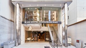 The exterior of And Hostel Shinsaibashi East with a modern design, including a glass entrance, a staircase, and a seating area outside.