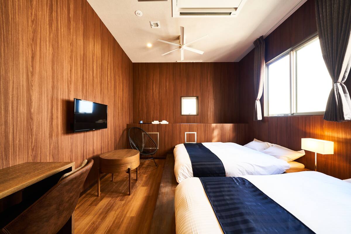 A hotel room with a wooden wall, a ceiling fan, a TV, a desk, a chair, two beds, and a window with curtains.