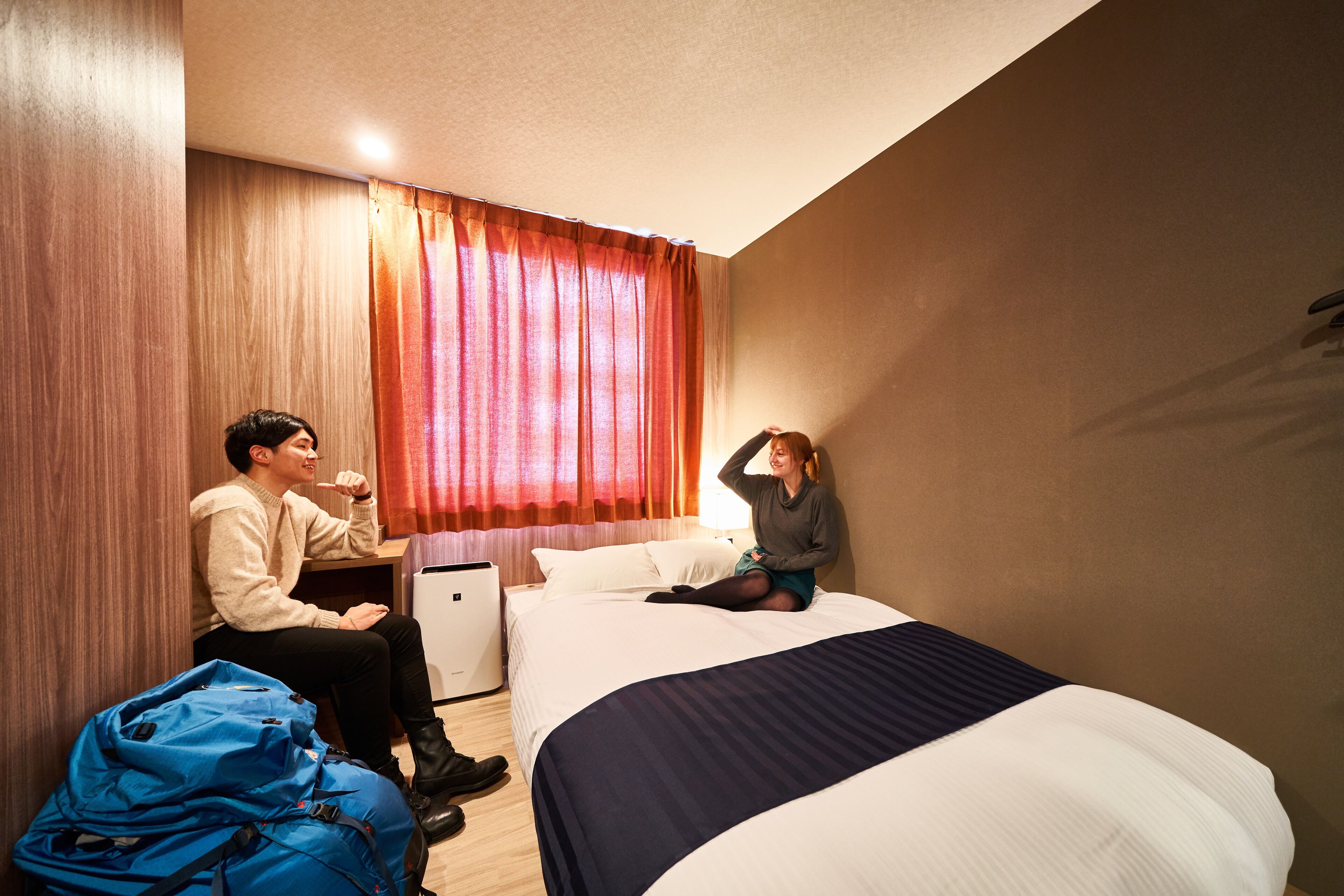 A hotel room with a bed, a desk, a chair, and a blue suitcase.