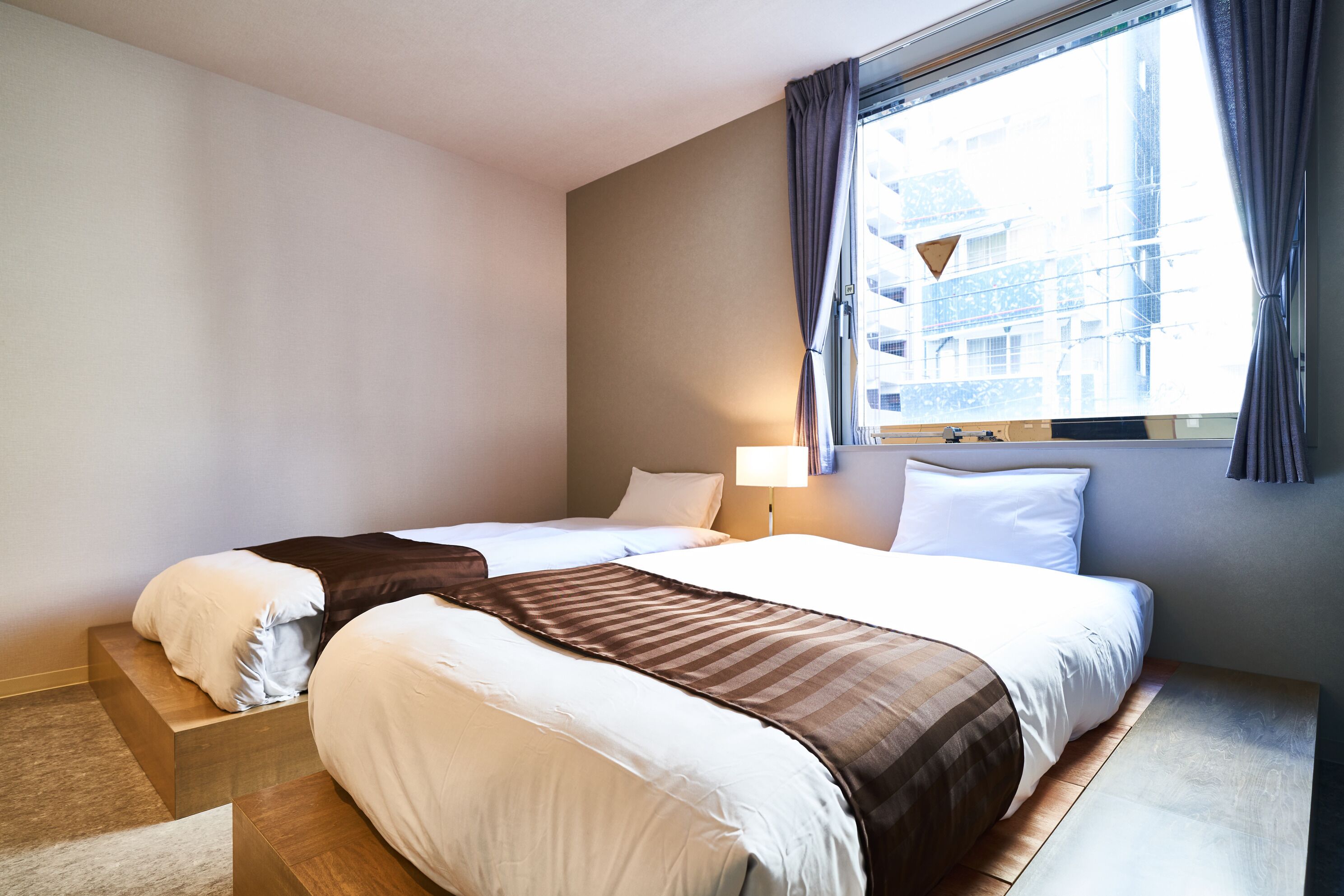 A hotel room with two beds, a window with curtains, a lamp, and a view of buildings outside.