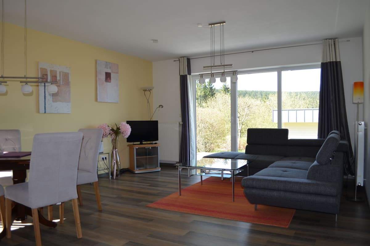 A modern living room with a sofa, a dining table with chairs, a TV, and a large window with curtains.
