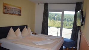 Un dormitorio con una cama grande, un cuadro en la pared, un televisor montado y un balcón con vistas a árboles.