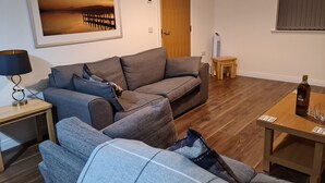A living room with a grey sofa, a small table with a bottle and glasses, a lamp, a framed picture on the wall, and a wooden door. - Linn of Avon Holiday Cottages (Ballindalloch)