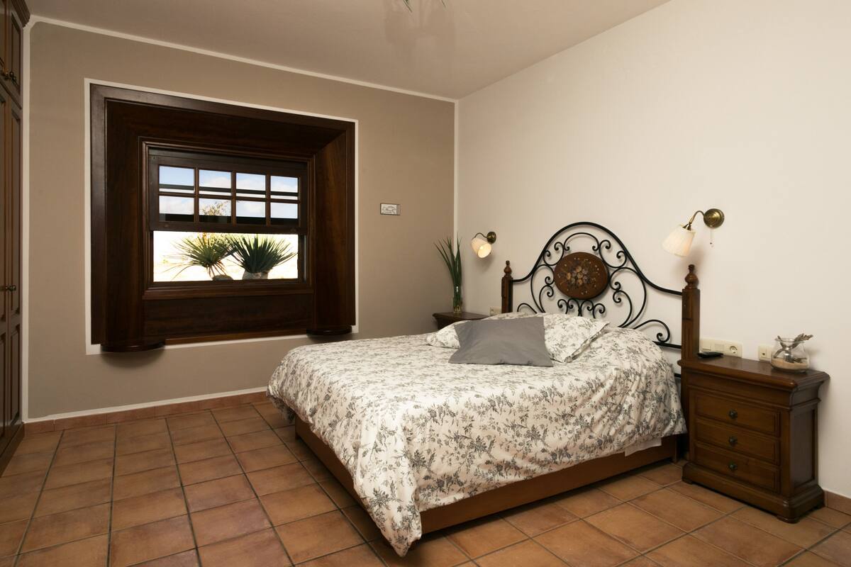 Quarto com cama, mesa de cabeceira, abajur, janela com vista para plantas e piso de azulejos.