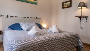 Una cama bien tendida con ropa de cama estampada, un cuadro de paisaje urbano en la pared y una lámpara de pared.