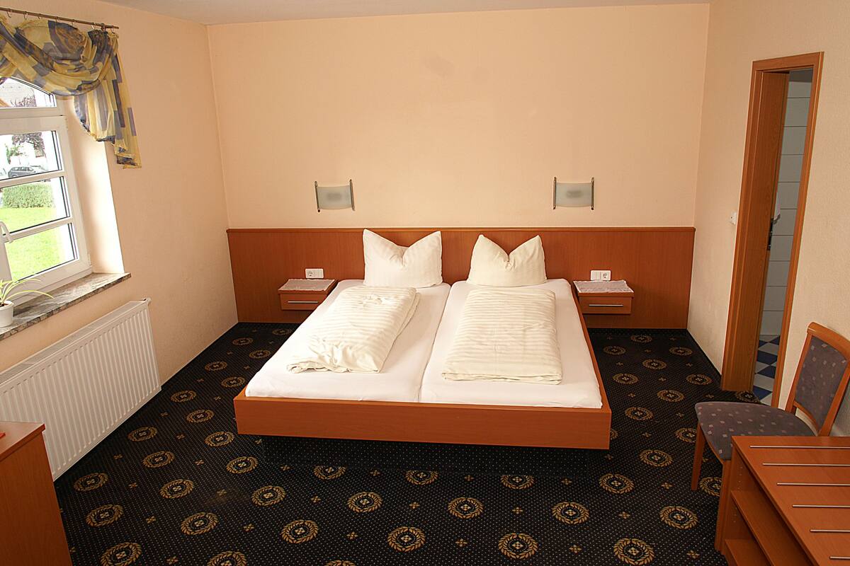 Habitación de hotel con una cama doble, dos mesitas de noche, una ventana con cortinas, un radiador, alfombra estampada y una silla de madera.