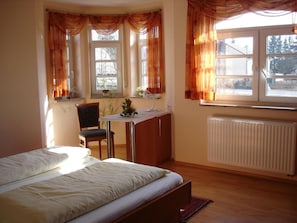 Ett sovrum med en säng, ett skrivbord och en stol. Det finns fönster med gardiner, ett element och utsikt över omgivningen.