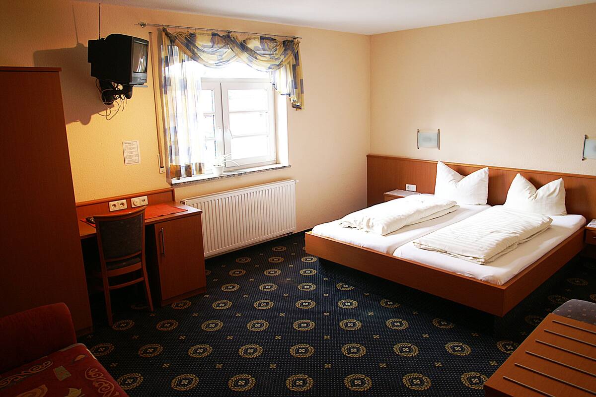 Habitación de hotel con cama, escritorio, silla, televisor montado en la pared y ventana con cortinas.