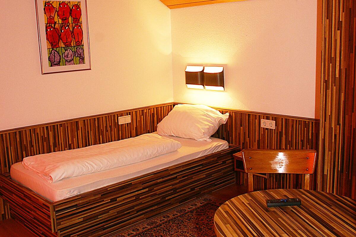 Habitación compacta con una cama individual, una pequeña mesa y una lámpara fijada a la pared.