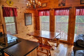 A wooden cabin interior with a dining table, benches, a chandelier, and windows with blinds.