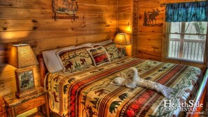 A cabin with a wooden interior, a bed with animal-themed bedding, and a window with a view of trees. - Game Room, Hot Tub, Wooded Views – Sleeps 12 – Near Dollywood & Pigeon Forge (Sevierville)