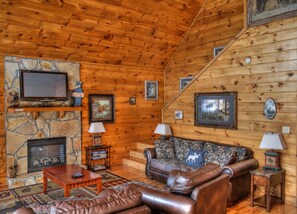 A cozy cabin interior with a stone fireplace, leather sofas, a television, and various framed pictures on the walls.