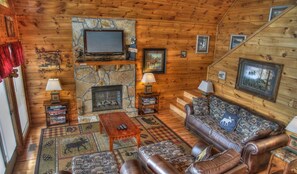 A cabin with a stone fireplace, leather furniture, and a television mounted on the wall. - Game Room, Hot Tub, Wooded Views – Sleeps 12 – Near Dollywood & Pigeon Forge (Sevierville)