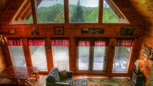 A cozy cabin interior with a wooden interior, large windows, a dining table, and a sofa. - Game Room, Hot Tub, Wooded Views – Sleeps 12 – Near Dollywood & Pigeon Forge (Sevierville)