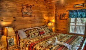 A cabin with a wooden interior, a bed with animal-themed bedding, and a window with a view of trees. - Game Room, Hot Tub, Wooded Views – Sleeps 12 – Near Dollywood & Pigeon Forge (Sevierville)