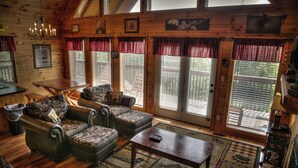 A cozy cabin interior with wooden walls, a dining area, and large windows with red curtains. - Game Room, Hot Tub, Wooded Views – Sleeps 12 – Near Dollywood & Pigeon Forge (Sevierville)