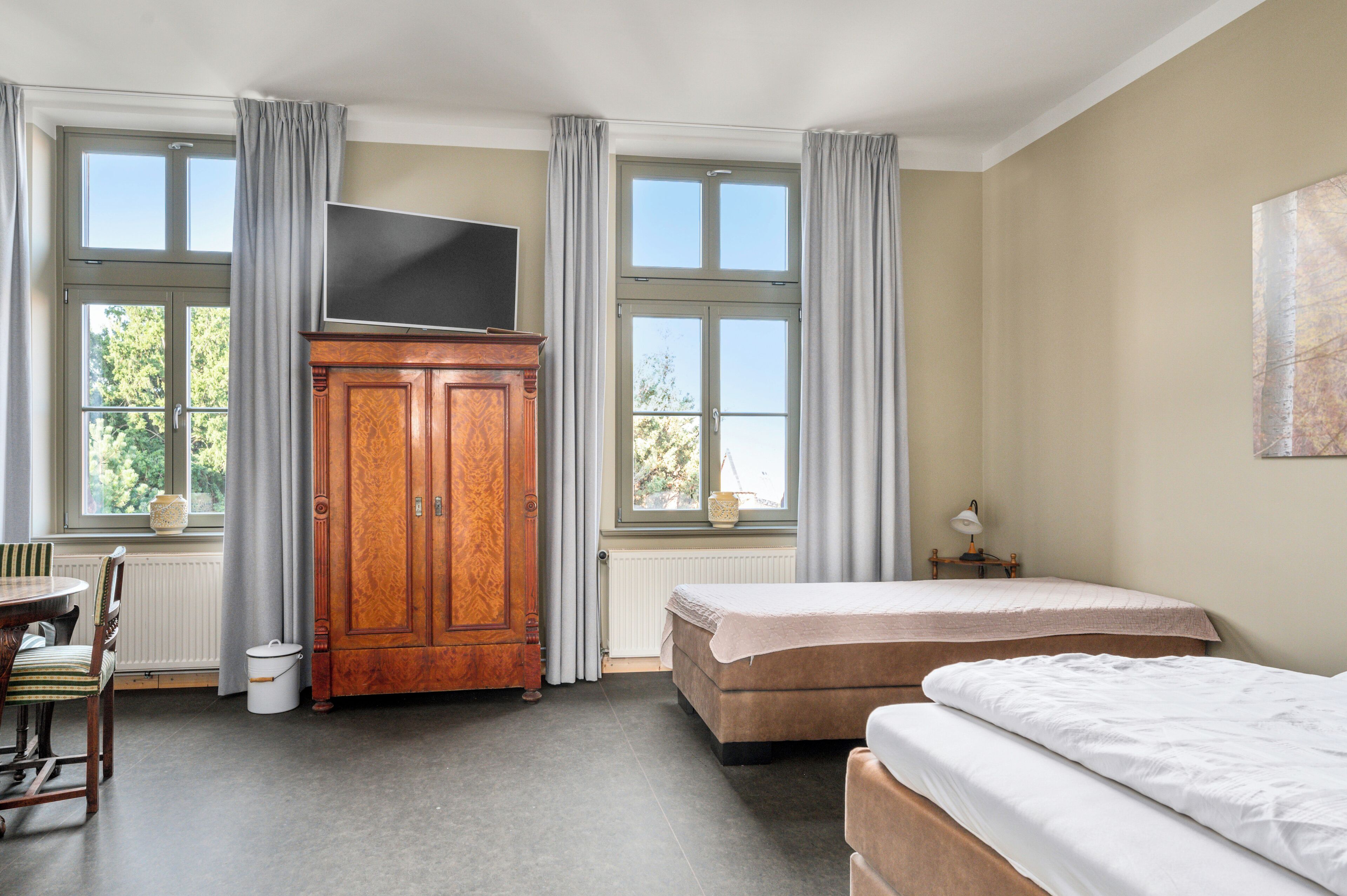 Ett hotellrum med två sängar, en garderob i trä, en tv, ett bord med stolar och stora fönster med gardiner.