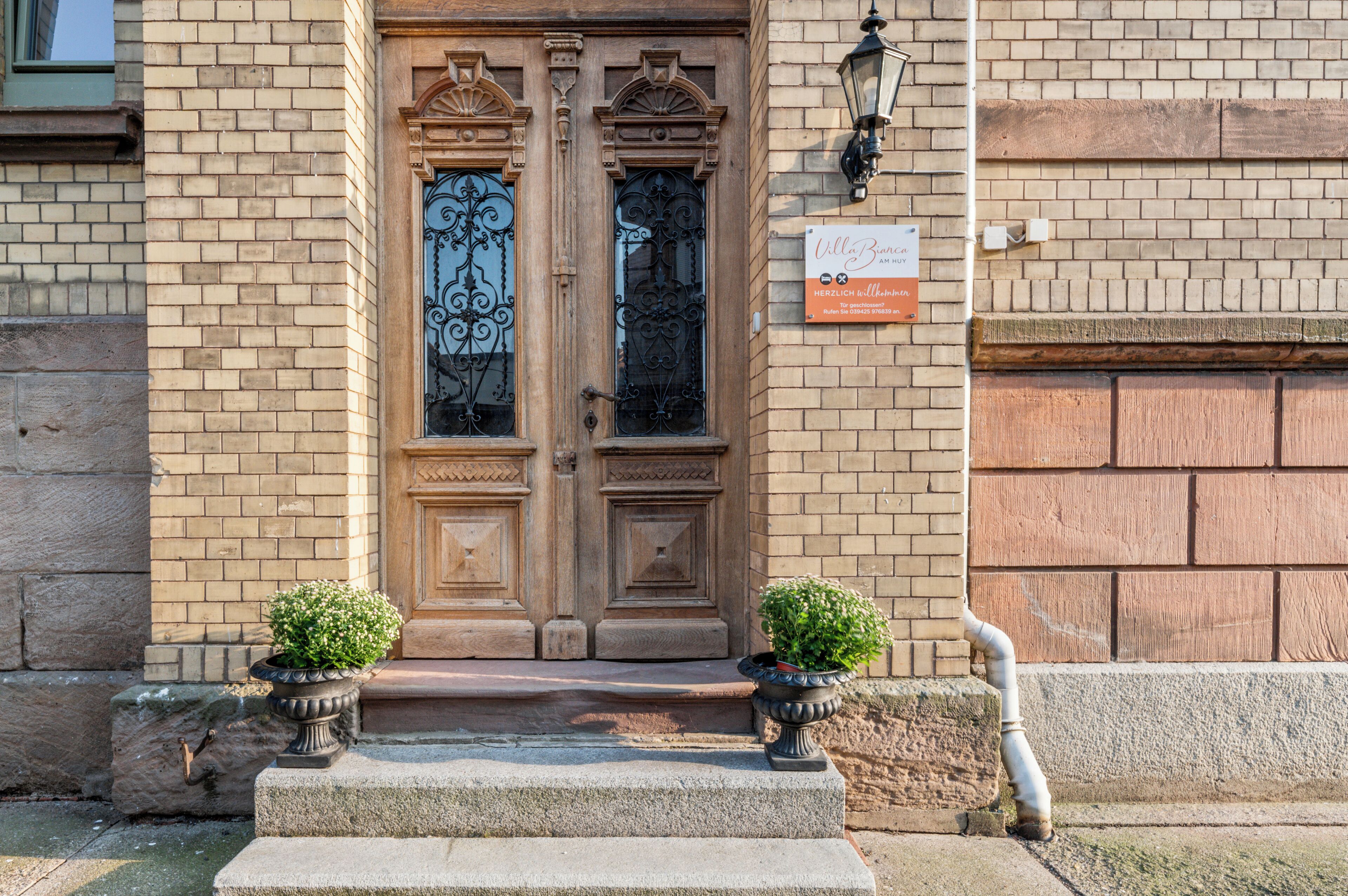 A building facade with a double wooden door, two potted plants on the steps, and a sign above the door.
